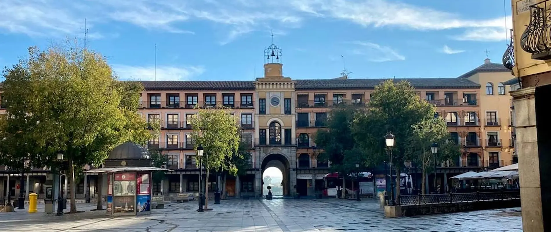 Plaza con edificios antiguos, árboles y un arco central; cielo azul y pavimento de piedra.