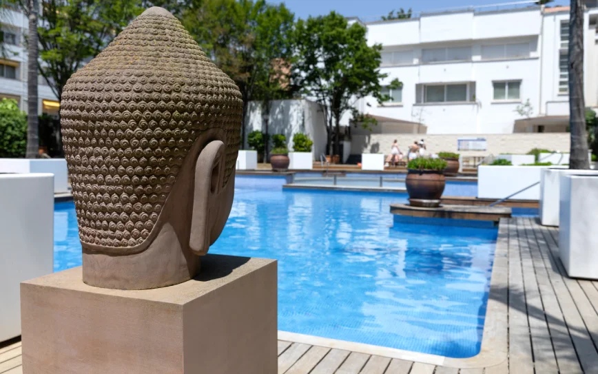 Con un busto zen en primer plano, de fondo la piscina. Todo con un aire cálido y relajante