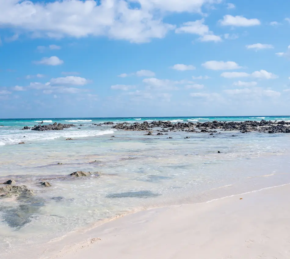 Playa con arena blanca, rocas dispersas, agua cristalina y cielo azul con nubes esponjosas.