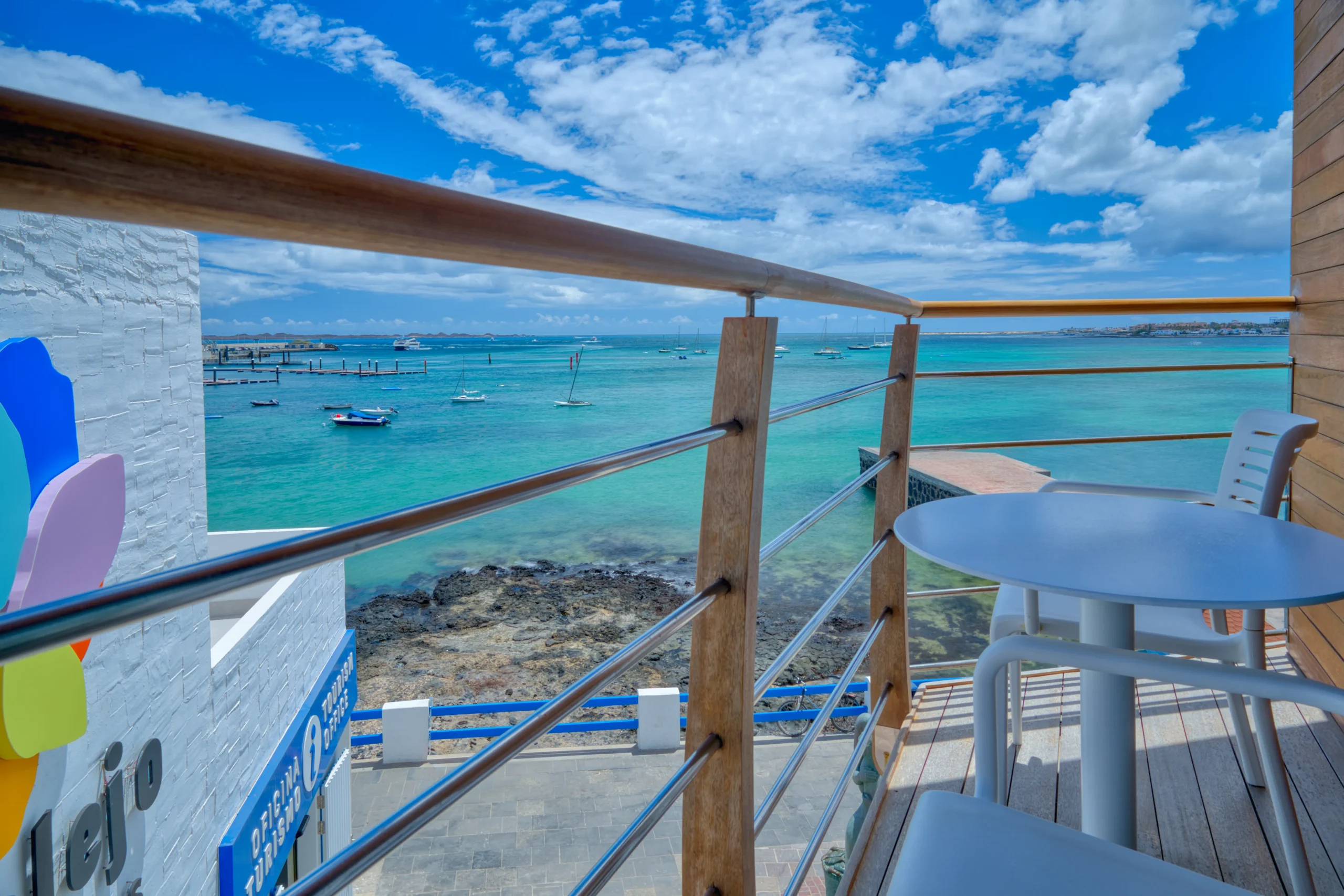 Vista desde un balcón hacia el mar turquesa, barcos pequeños y cielo azul con nubes. Mobiliario blanco en primer plano.