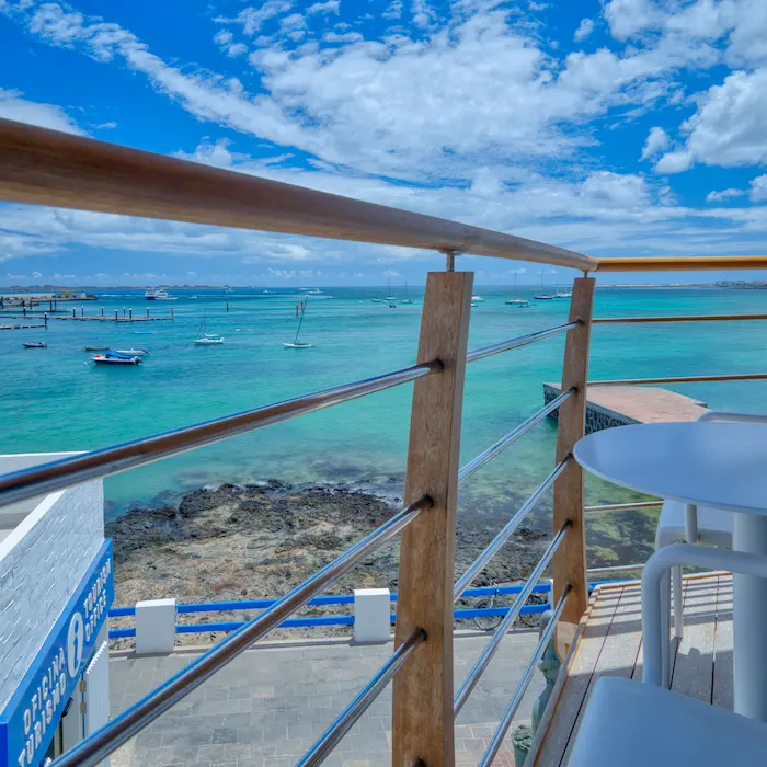 Vista desde un balcón con baranda de madera hacia un mar turquesa y cielo azul. Hay una mesa blanca y barcos lejanos.