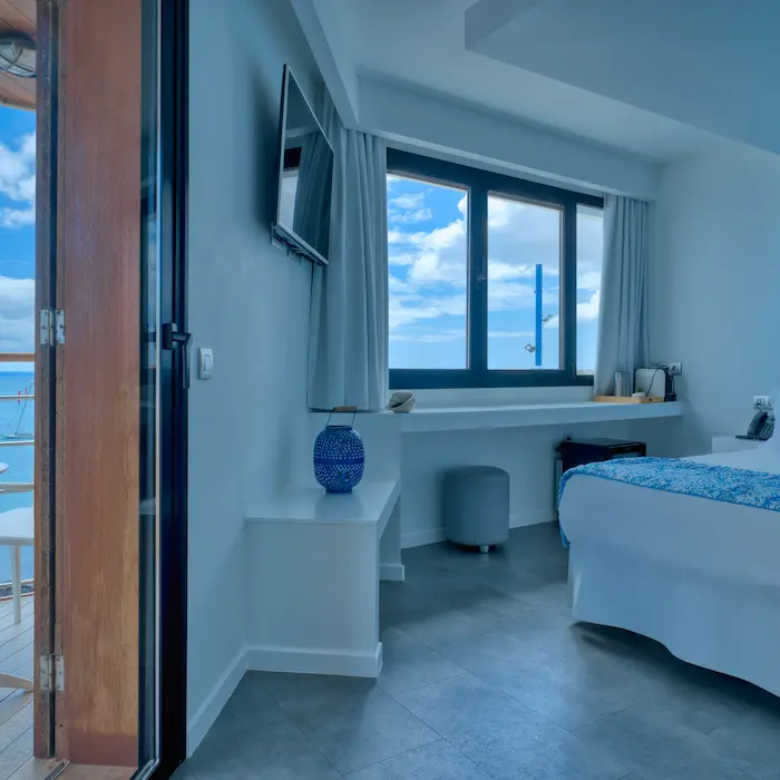 Habitación moderna con ventana grande, cama, televisor, lámpara azul y vistas al mar y cielo azul con nubes.