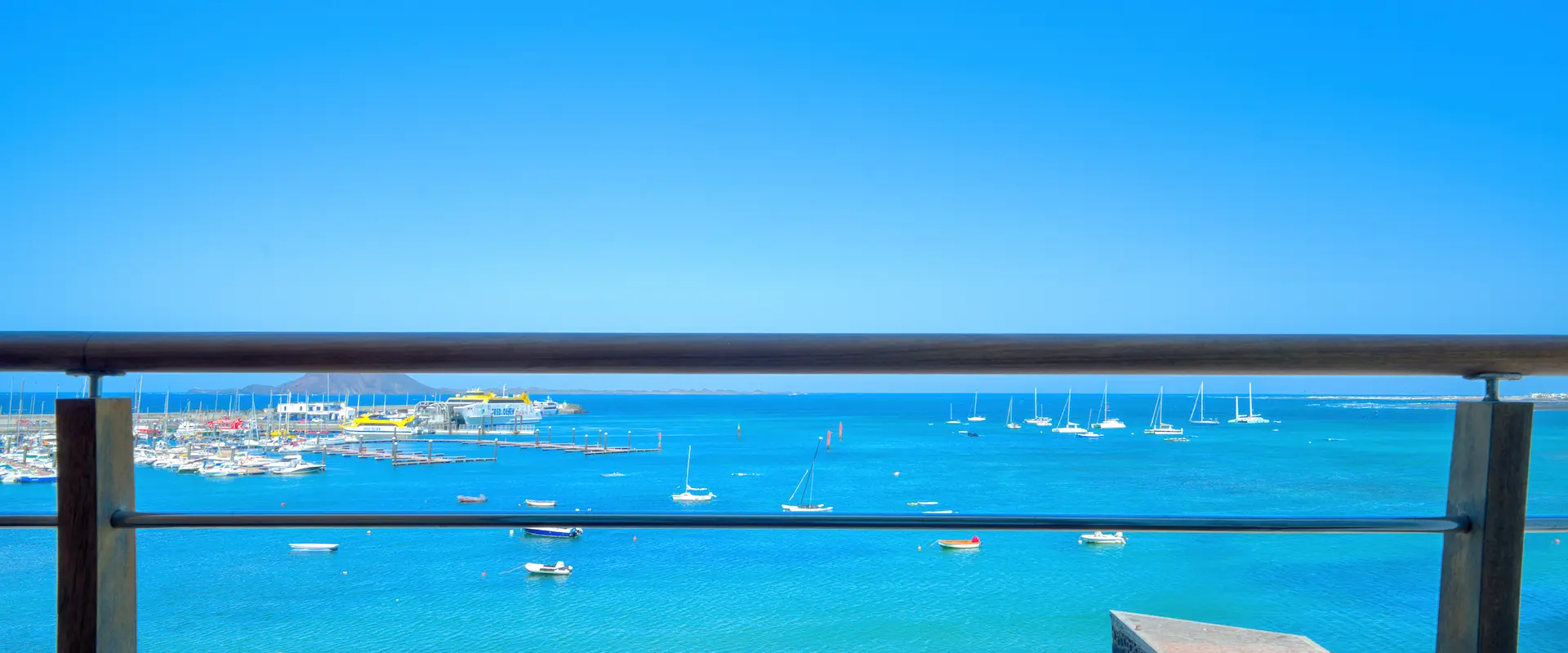 Vista de un puerto con barcos en aguas azules bajo un cielo despejado, vista desde una barandilla.