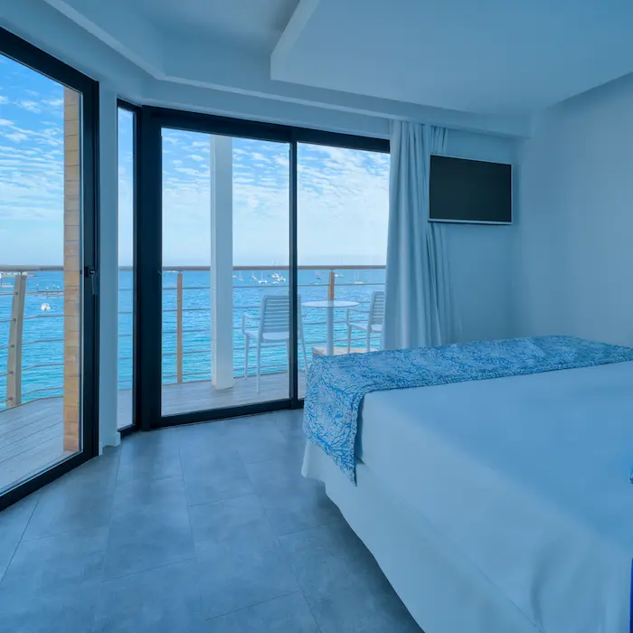 Habitación de hotel con cama y balcón con vista al mar, sillas blancas y televisor en la pared. Cielo despejado.