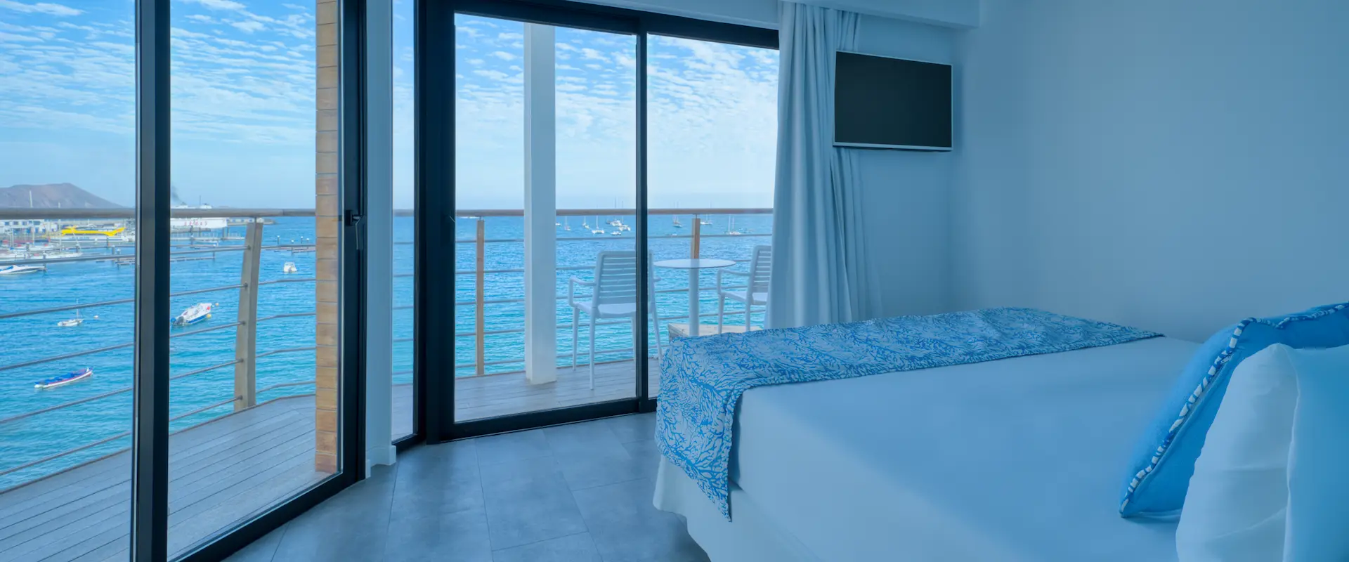 Habitación de hotel moderna con vista al mar, balcón, cama blanca y decoración en tonos azules.