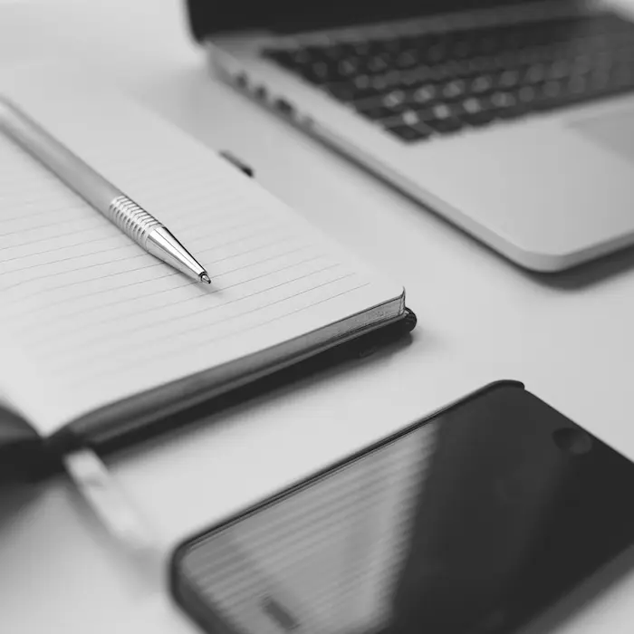 Cuaderno abierto con bolígrafo sobre líneas, junto a una parte de una laptop y un teléfono en tono monocromático.