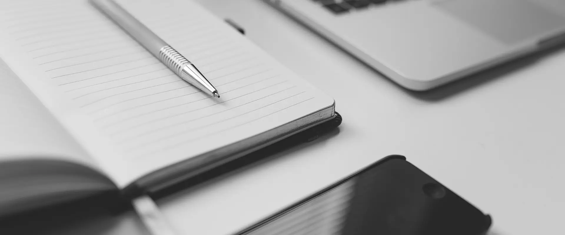 Cuaderno abierto con bolígrafo plateado, teléfono y laptop en blanco y negro sobre un escritorio.