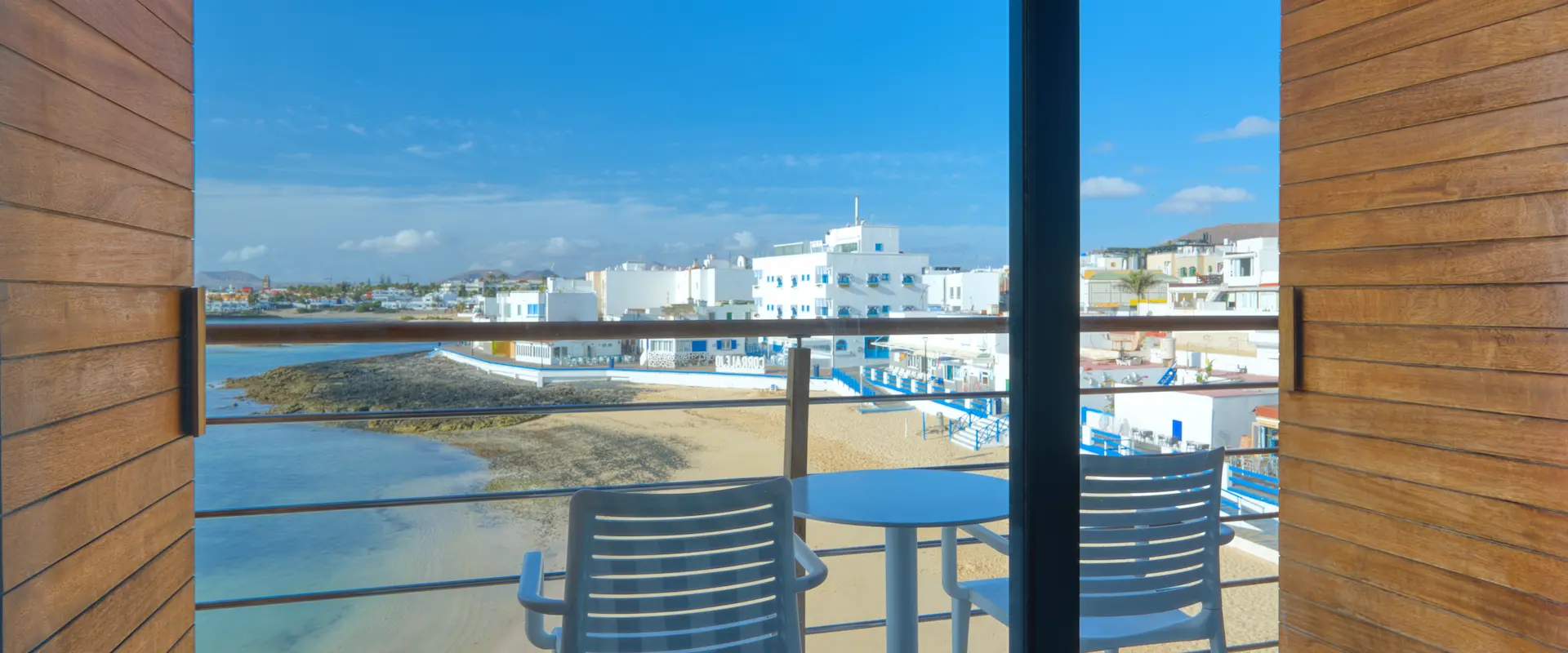 Vista desde un balcón con sillas y una mesa hacia una playa y edificios blancos bajo un cielo azul.