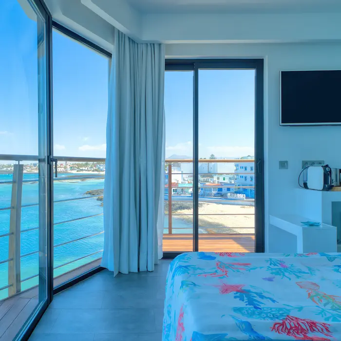 Habitación con vista al mar, cama colorida, ventanas amplias y TV en la pared. Balcón con barandal.