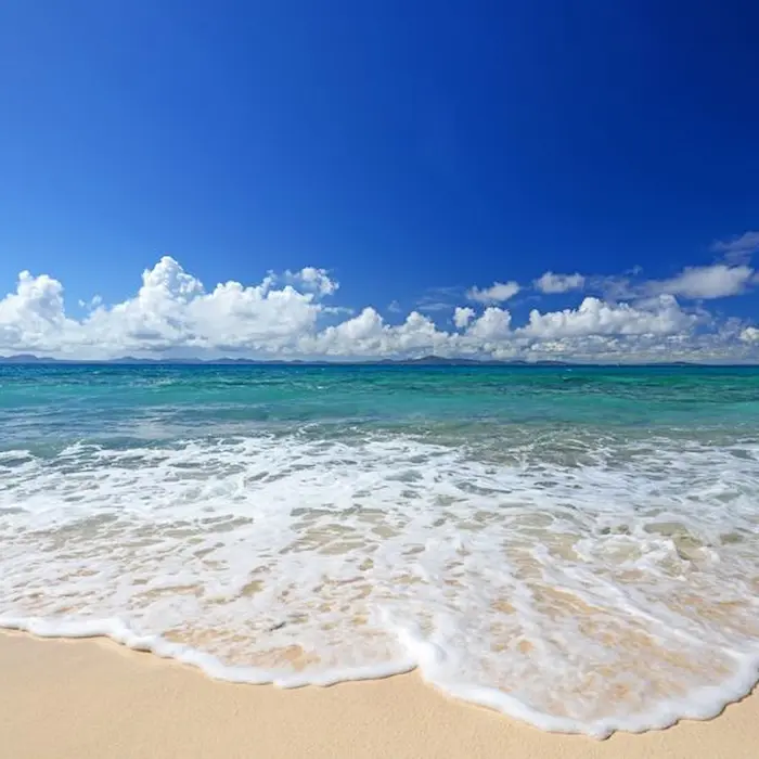 Playa con olas suaves, arena blanca y cielo azul con algunas nubes.