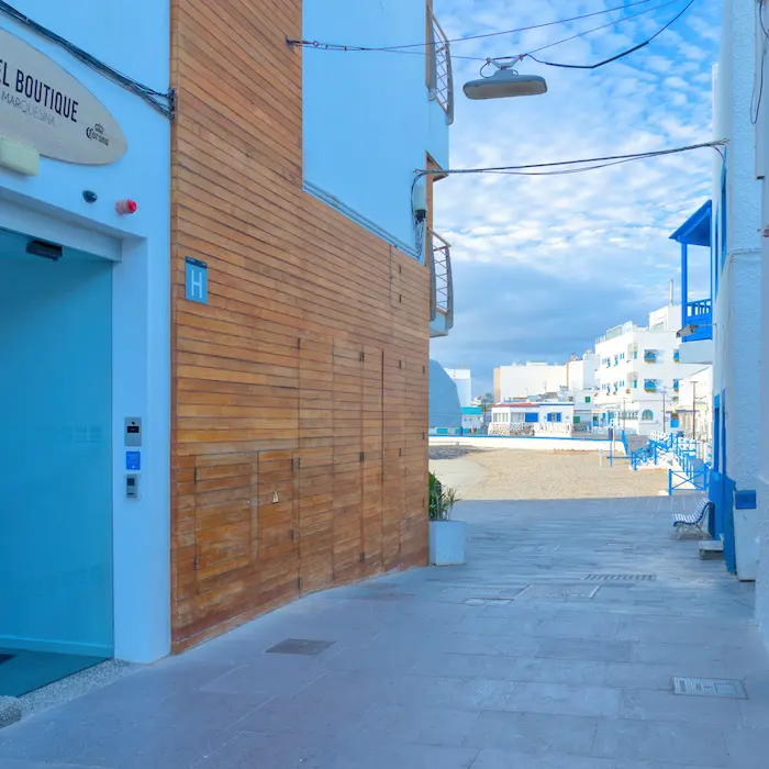 Calle estrecha entre edificios, uno con fachada de madera, que conduce a una playa con cielo parcialmente nublado.