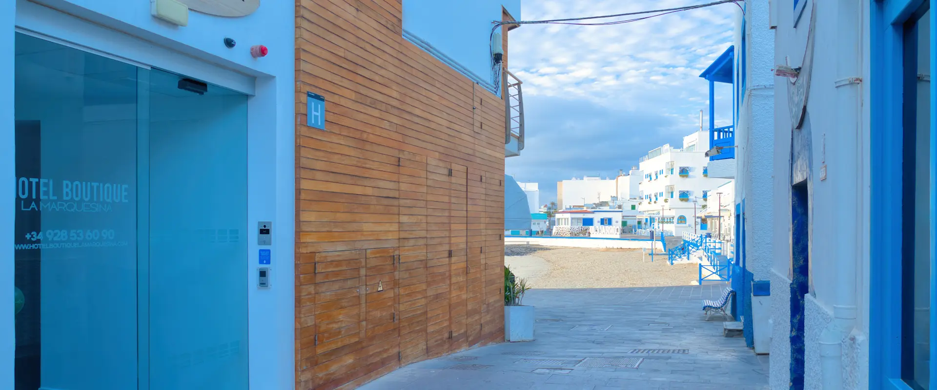 Calle estrecha con paredes de madera y un hotel a la izquierda, conduce a una playa con construcciones blancas y azules.