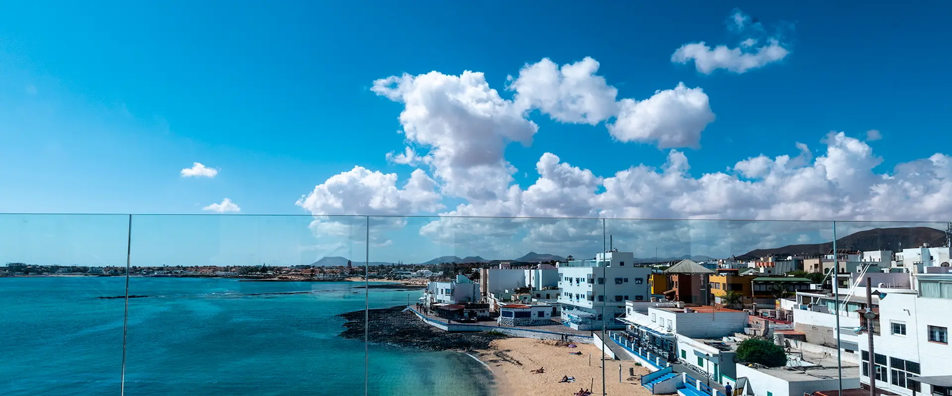 Vista panorámica de un pueblo costero con cielo azul, nubes esponjosas, playa y edificios junto al mar.