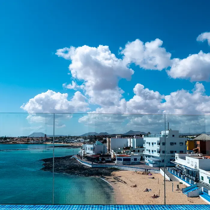Vista de una costa con edificios, cielo azul y nubes, desde un balcón con piscina y cristal transparente.
