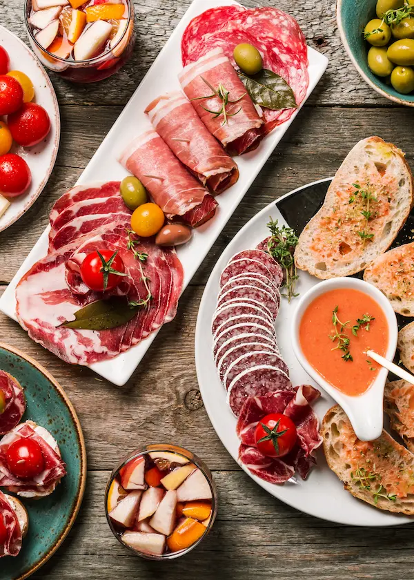 Tabla de embutidos variados con aceitunas y pan, cerca de vasos de sangría y un plato con tomate rallado.
