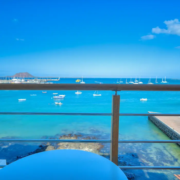 Vista desde balcón con barandas al mar azul y pequeños barcos; cielo despejado con pocas nubes.