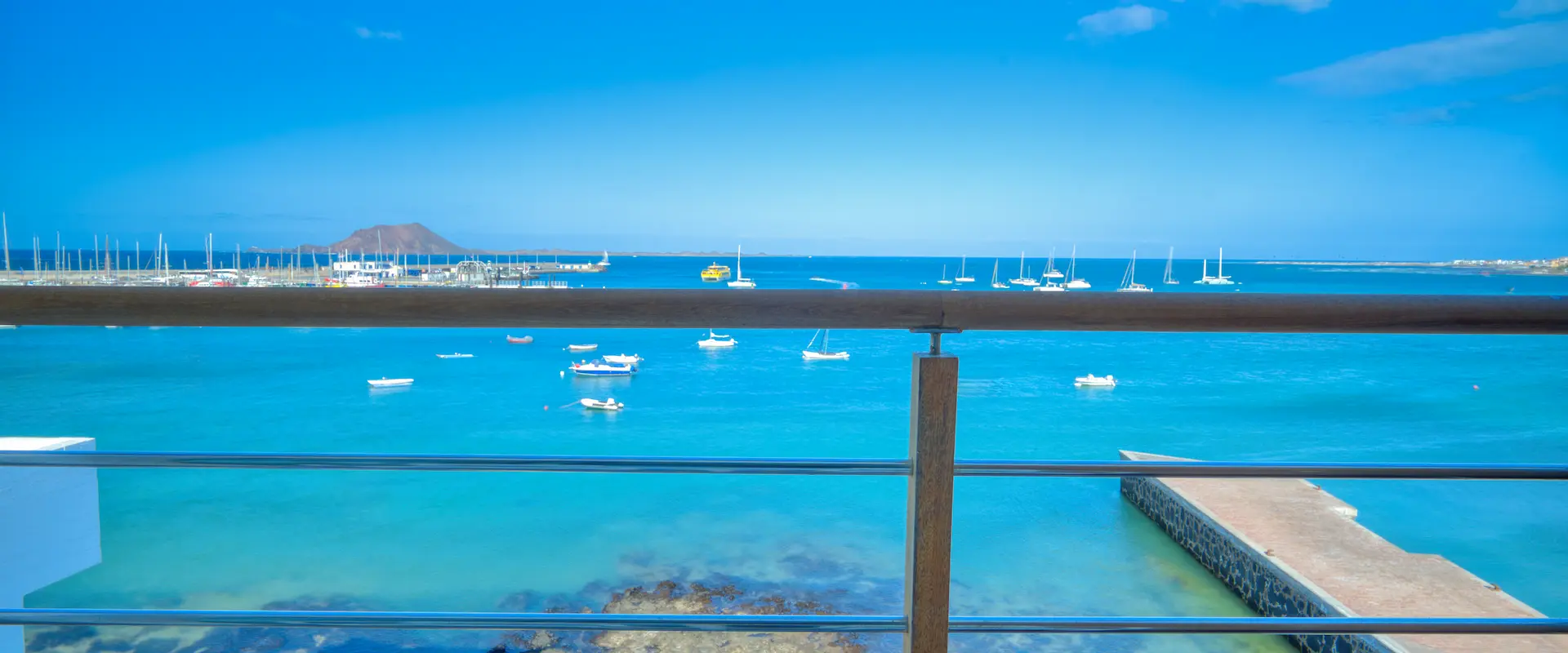 Vista al mar desde un balcón, con barcos en el agua y un cielo azul despejado.