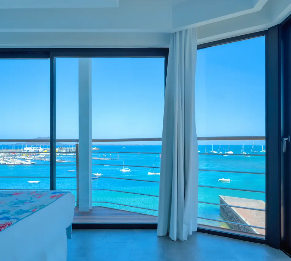 Vista desde una habitación con ventanales al mar, mostrando un puerto con barcos y un cielo despejado.