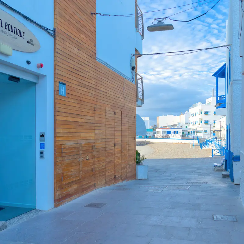 Pasillo entre edificios hacia la playa, cielo azul con nubes dispersas. Fachadas blancas y detalles en azul.