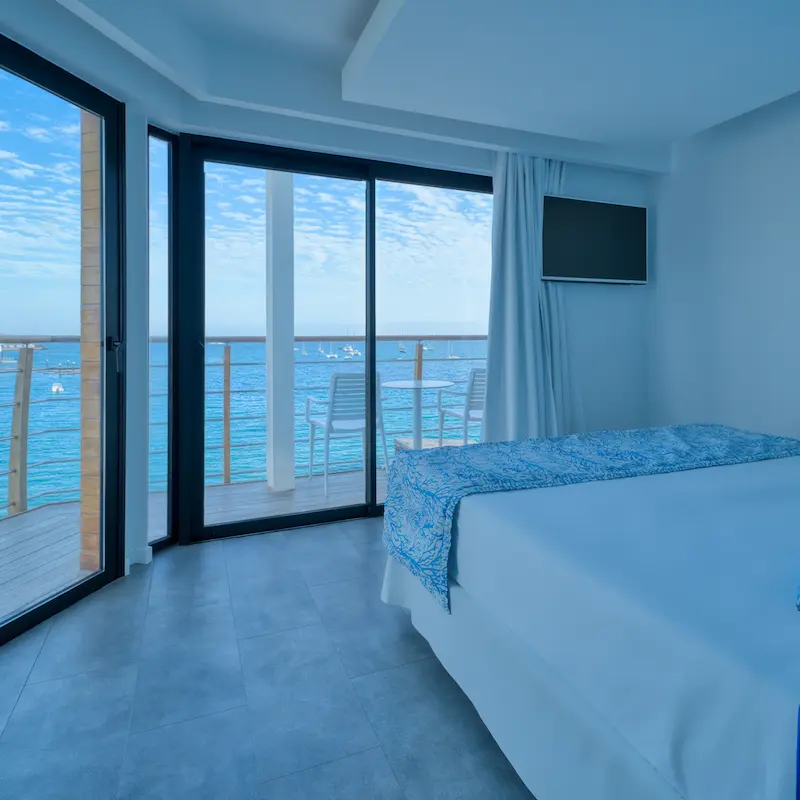 Habitación con vista al mar a través de ventanas grandes; balcón con sillas y mesa, cielo despejado.