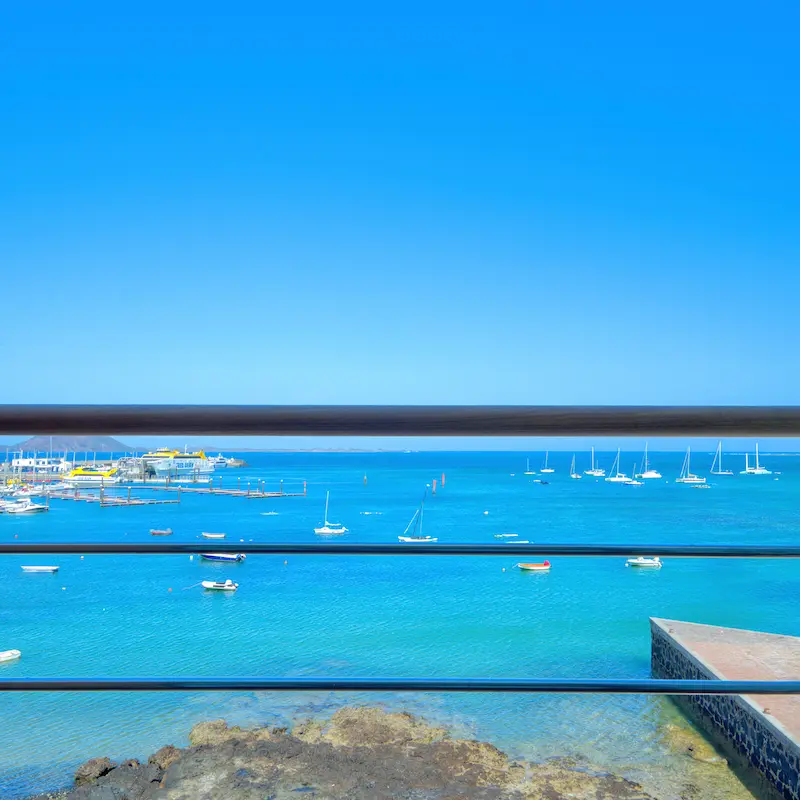 Vista al mar con barcos de vela anclados, cielo azul despejado y barandilla en primer plano.