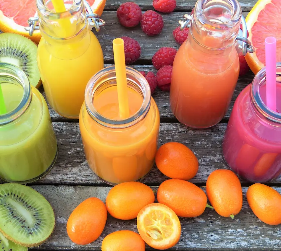 Botellas de jugo colorido con popotes en mesa de madera rodeadas de frutas como kiwi, kumquats, frambuesas y toronjas.