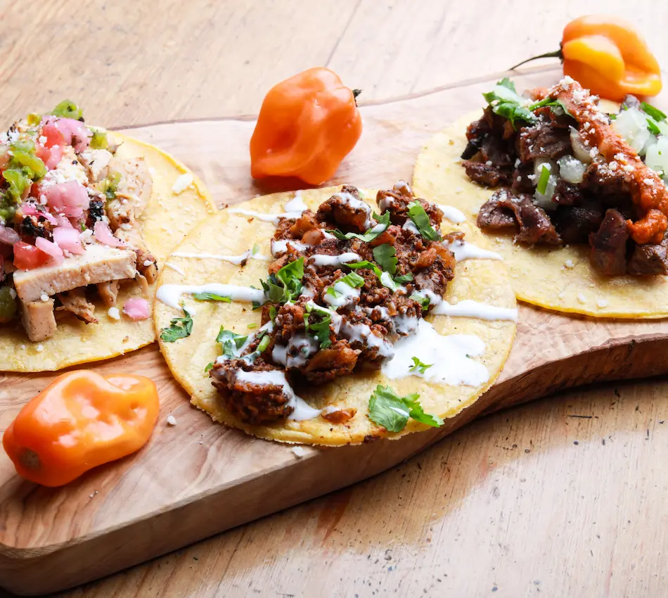 Tres tacos variados sobre tabla de madera con dos chiles habaneros al lado.