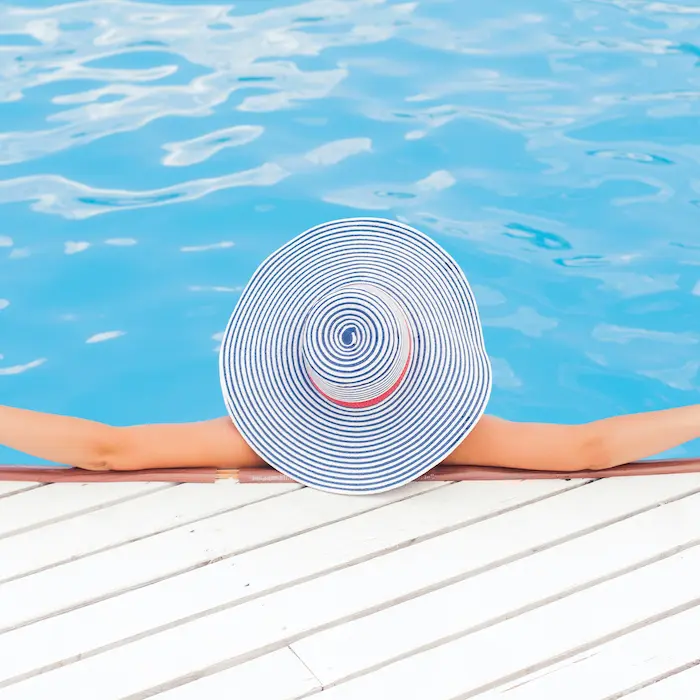 Persona con sombrero de rayas descansa al borde de una piscina azul brillante.