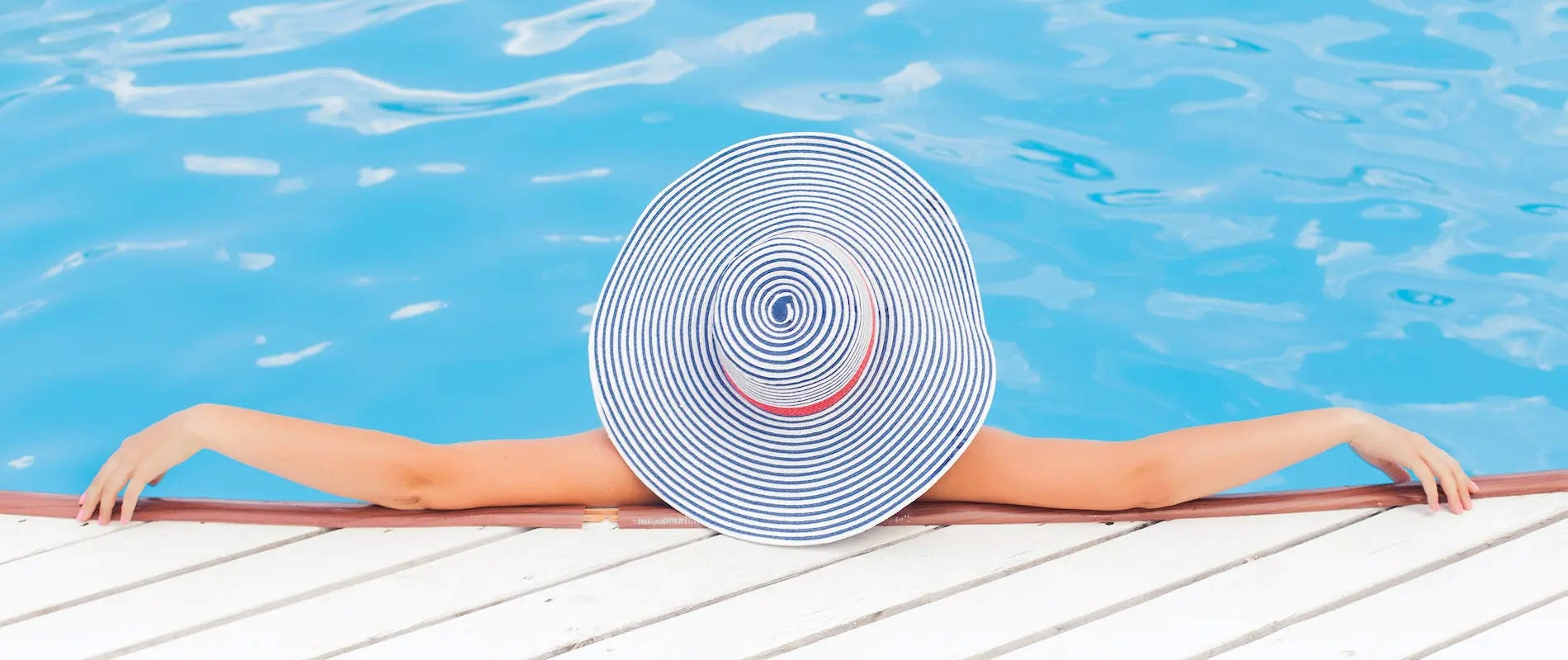 Persona con sombrero rayado se relaja en una piscina, apoyada en el borde de madera blanca.