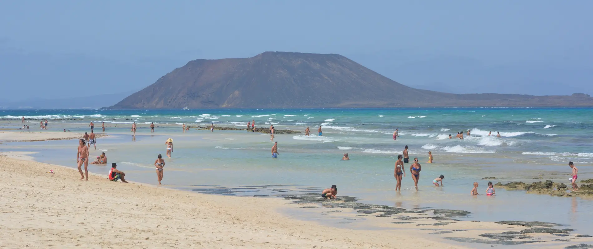 Personas disfrutando en una playa de aguas cristalinas, con una montaña al fondo y olas en el mar.