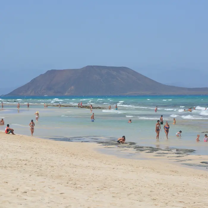 Personas disfrutando de una playa soleada con agua azul turquesa y una montaña en el fondo.