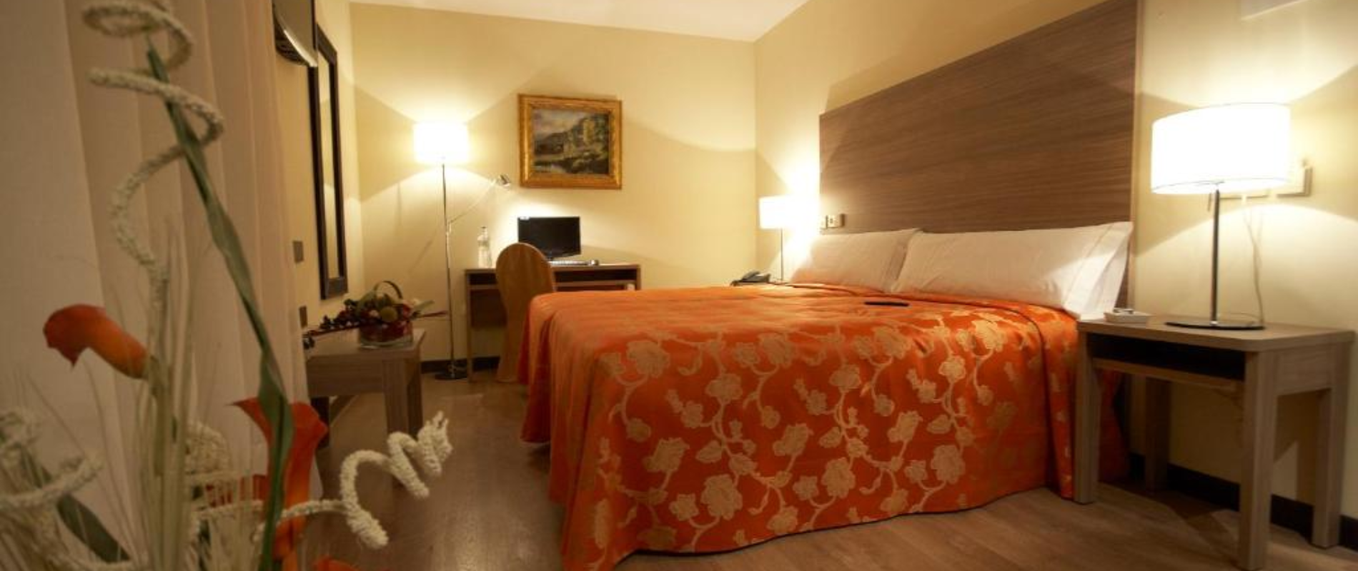 Habitación de hotel con cama grande, cubierta por colcha naranja, escritorio con silla y lámparas en mesitas.
