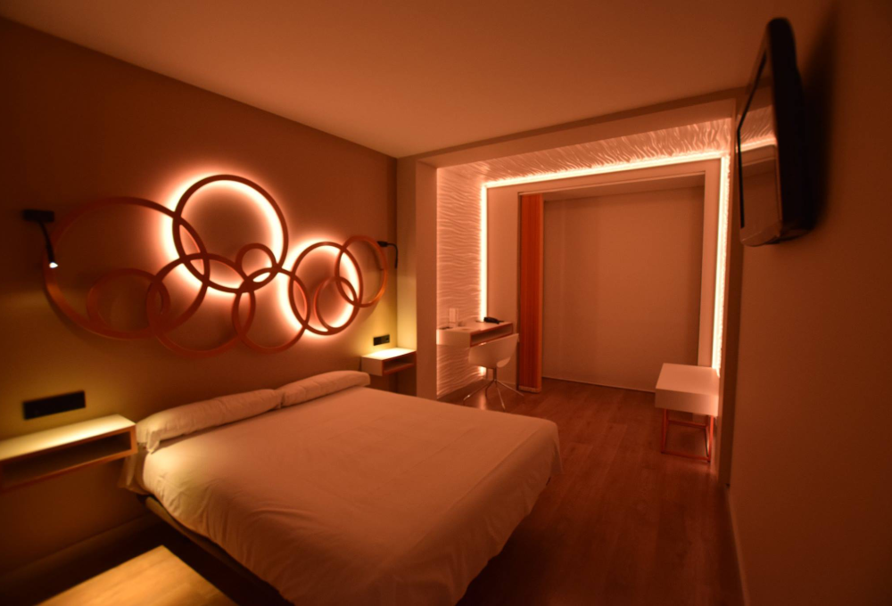 Habitación moderna con cama, decoración circular iluminada y escritorio. Luces cálidas crean ambiente acogedor.