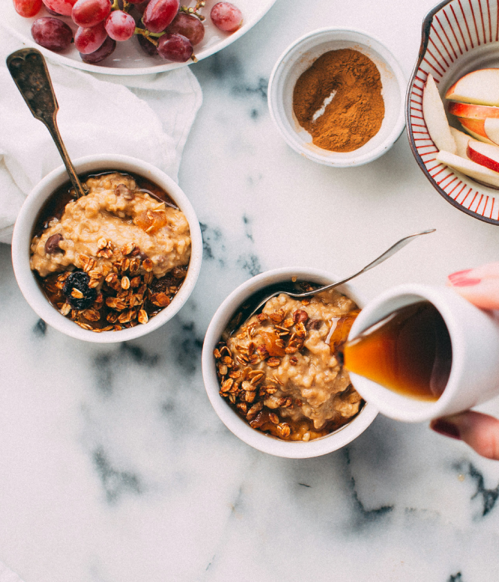 Dos tazones de avena con frutas y granola, jarabe siendo vertido, acompañados de uvas, canela y rodajas de manzana.
