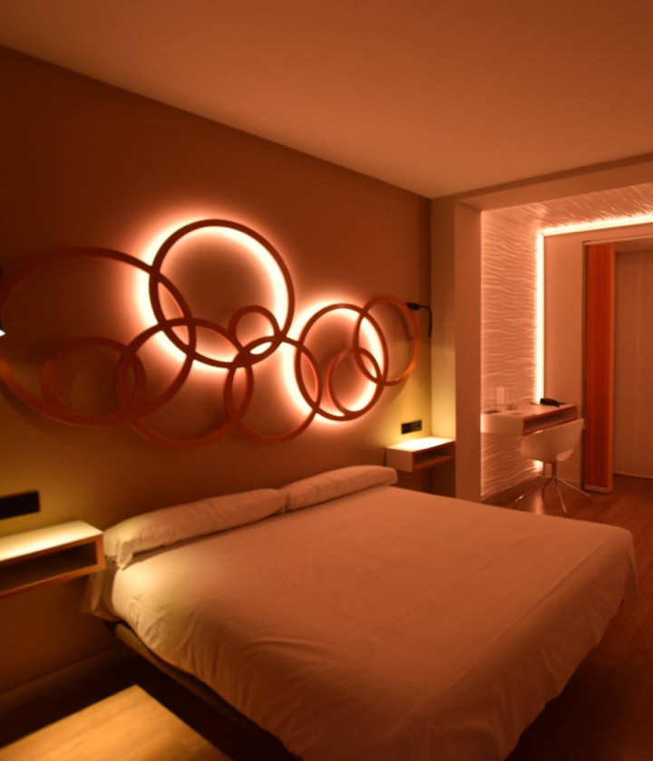 Habitación moderna con cama doble y obra de arte circular iluminada en la pared. Mesas laterales y escritorio.