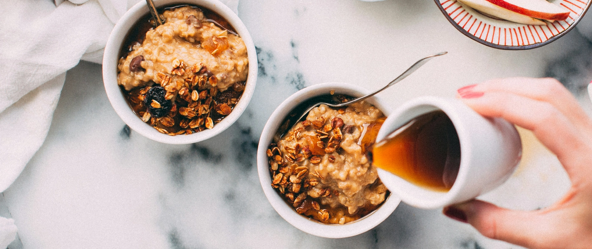 Dos tazones de avena con granola y uvas pasas, mientras se vierte miel o jarabe sobre uno de ellos.