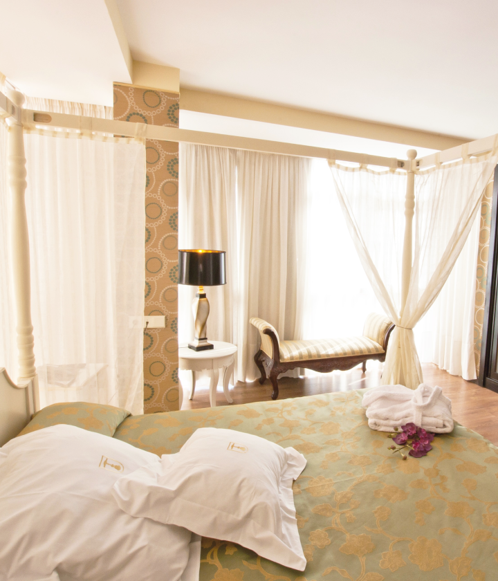 Habitación luminosa con cama cubierta de almohadas, dosel, lámpara de mesa y chaise longue junto a una ventana amplia.
