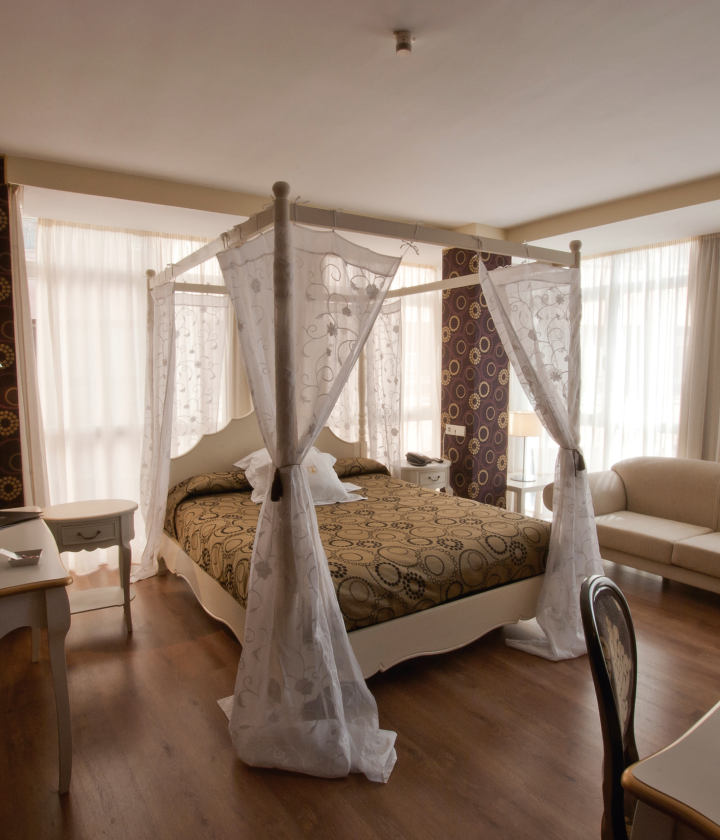 Habitación con cama con dosel, cortinas blancas, decoración clásica y ventanales con mucha luz natural.