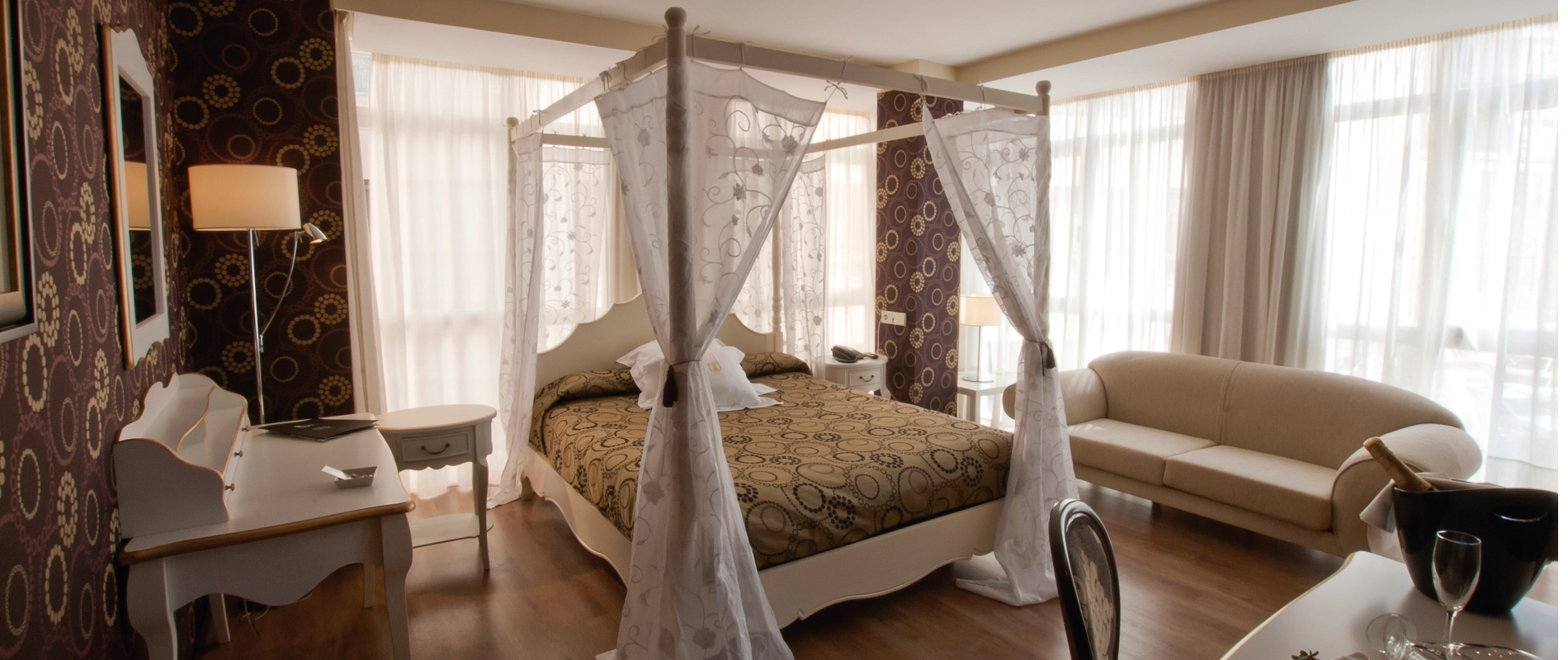 Habitación elegante con cama con dosel, papel tapiz decorativo, sofá beige y luz suave entrando por cortinas blancas.