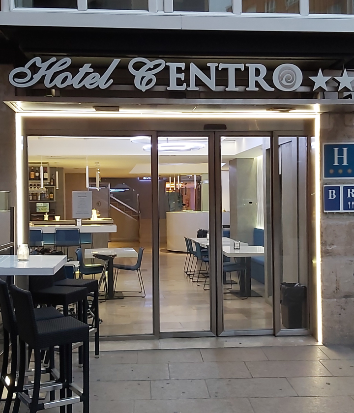 Entrada de hotel moderno con mesas y sillas en el interior y luces brillantes alrededor.