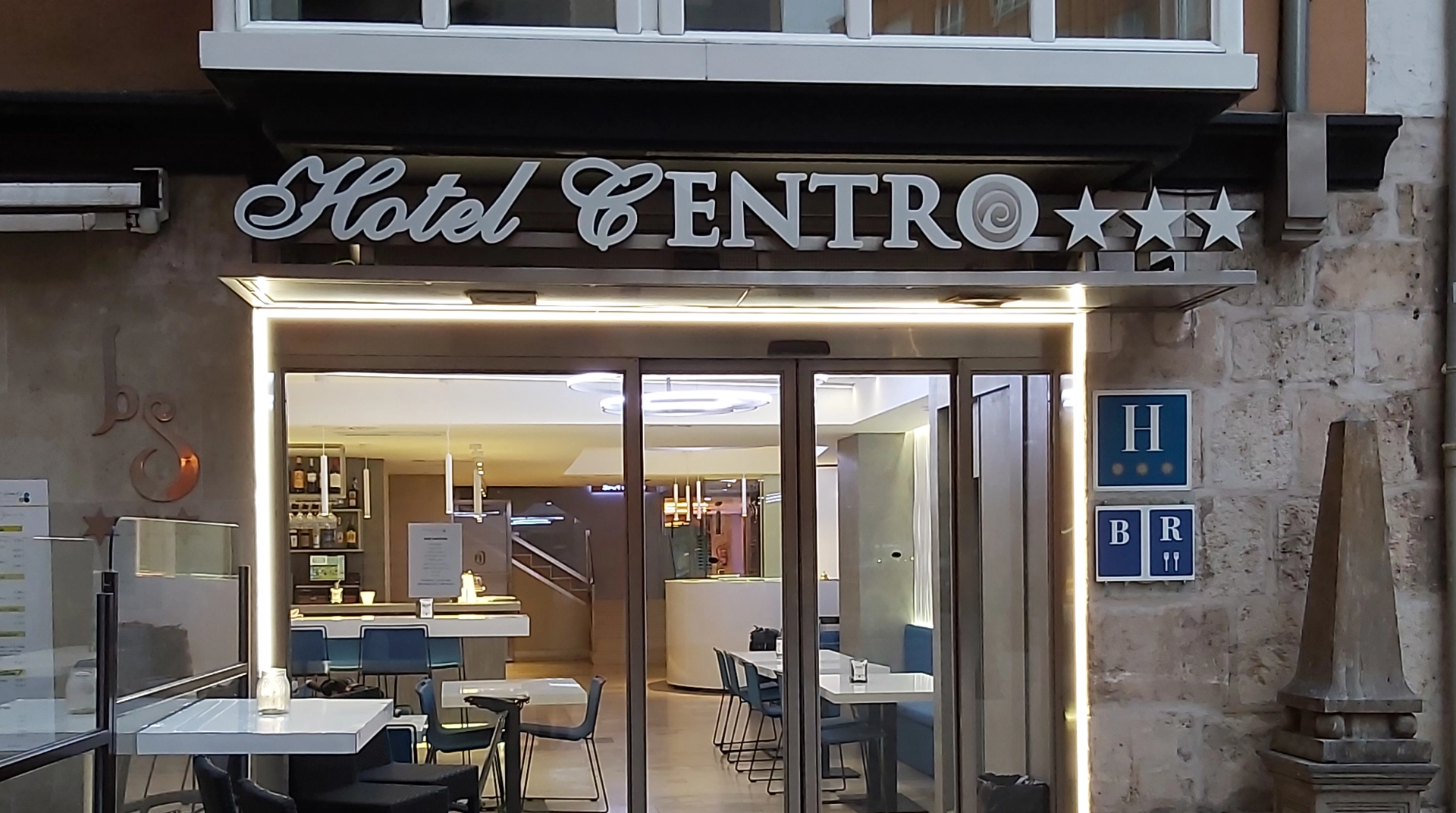 Entrada iluminada del Hotel Centro de tres estrellas, con sillas y mesas en el interior visible a través del cristal.