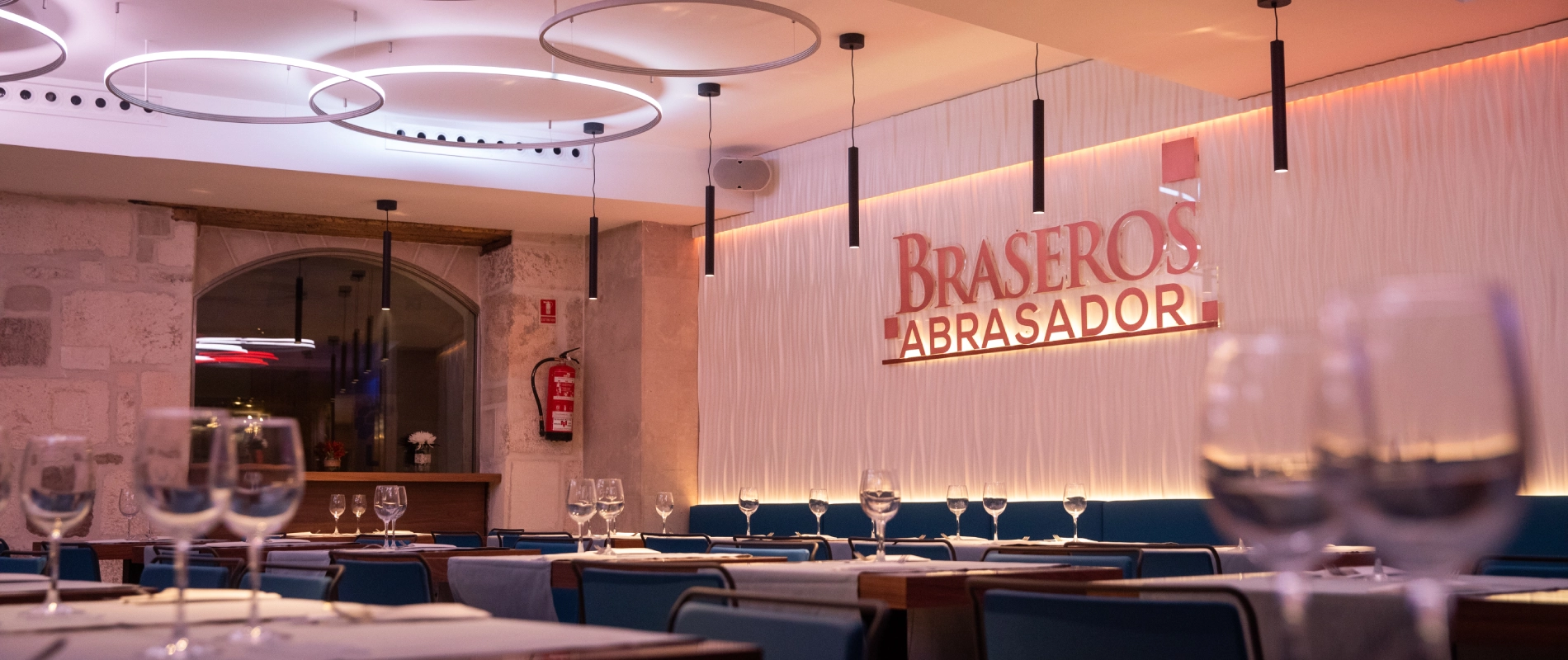 Restaurante elegante con mesas puestas y copas de vino, iluminación moderna y un cartel que dice "BRASEROS ABRASADOR".
