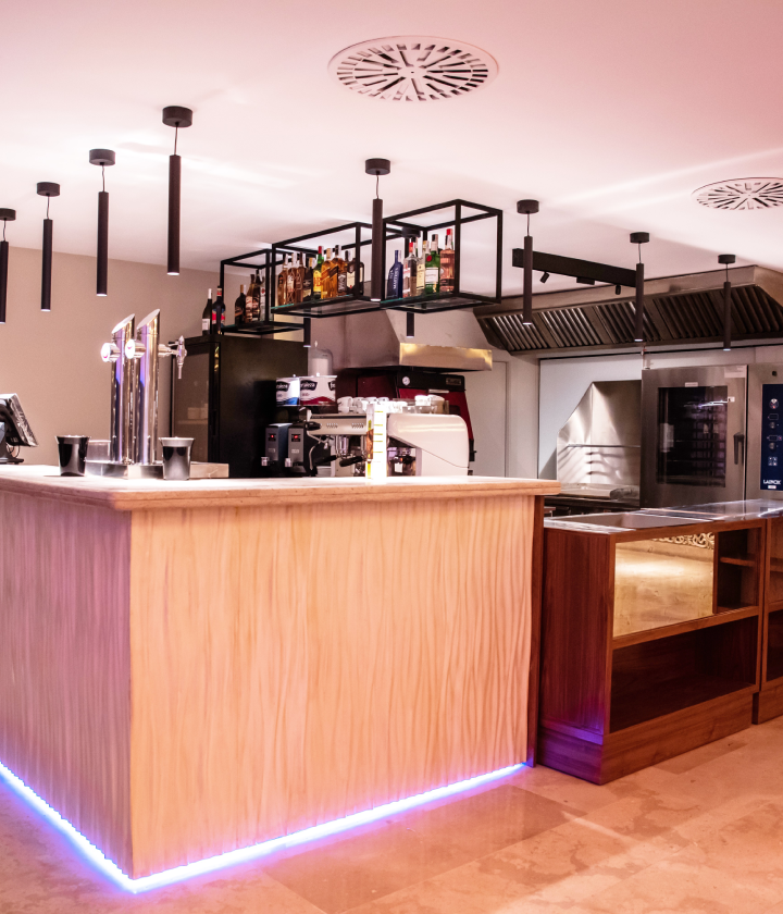 Bar moderno con luces colgantes, estantes de licor, dispensadores de cerveza y cafeteras sobre la barra.