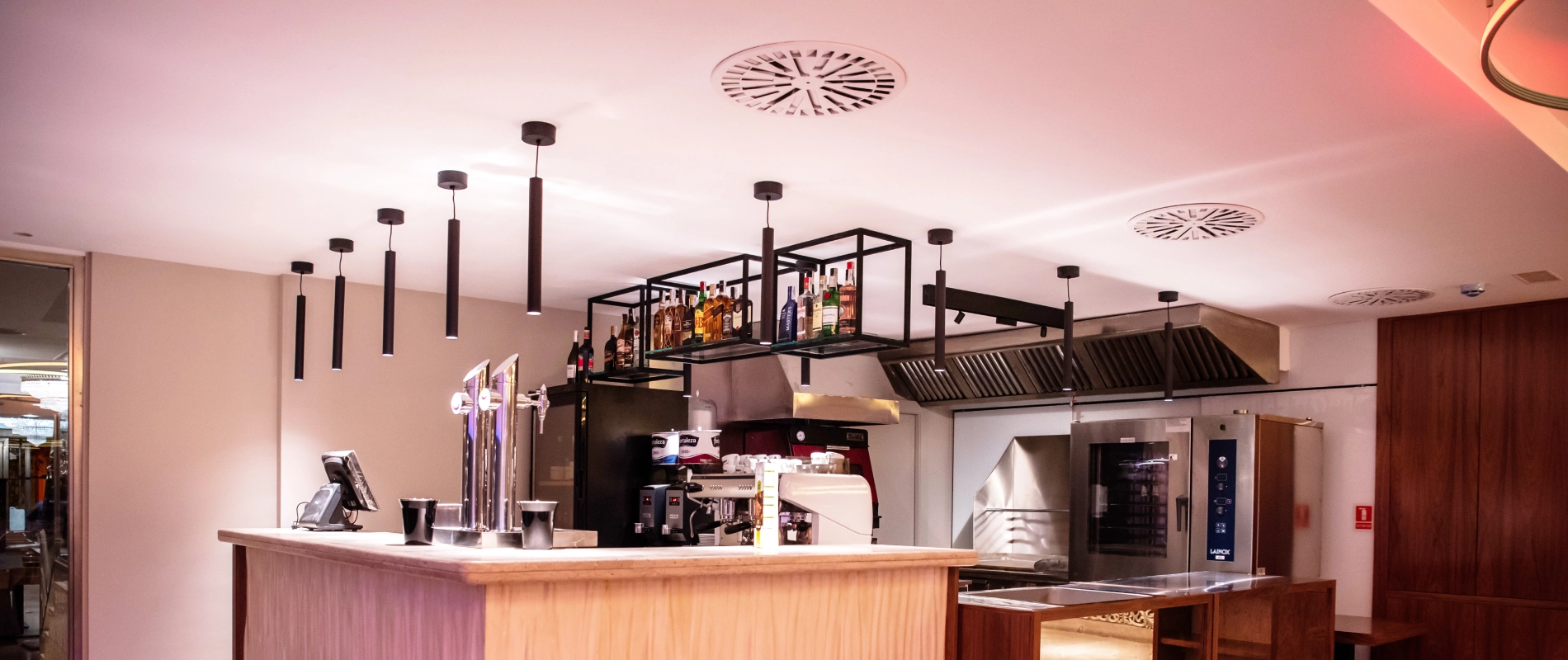 Barra de bar moderna con botellas en estantería arriba, luces colgantes y máquinas de café bajo ventilación.