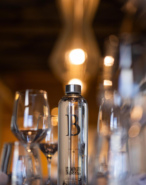 Botella de agua entre copas de vino, con una lámpara encendida en el fondo borroso.