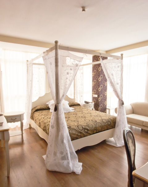 Dormitorio elegante con cama con dosel, cortinas blancas, fondo con papel tapiz decorativo y sillón beige.