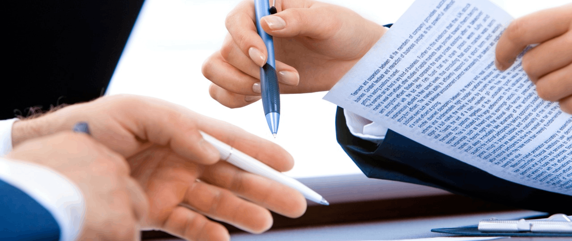 Manos revisando un documento en papel, sosteniendo bolígrafos, en un entorno de oficina.