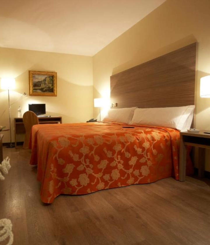 Habitación de hotel con cama doble, edredón naranja, escritorio con silla, lámparas y cuadro en la pared.