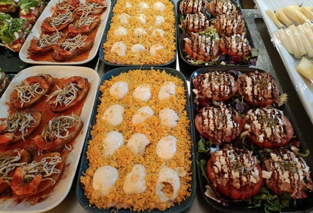 Variedad de platos fríos: tostadas con mariscos, ensalada de huevo y tomate con aderezo, dispuestos en bandejas.