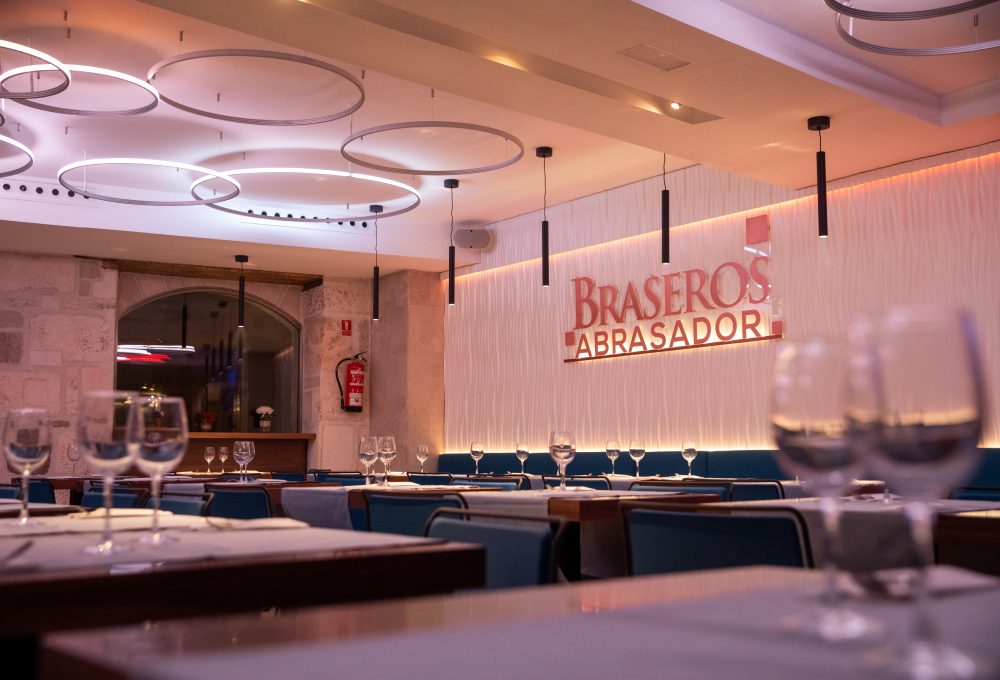 Interior de un restaurante elegante con mesas listas y decoración moderna. Un letrero dice "Braseros Abrasador".
